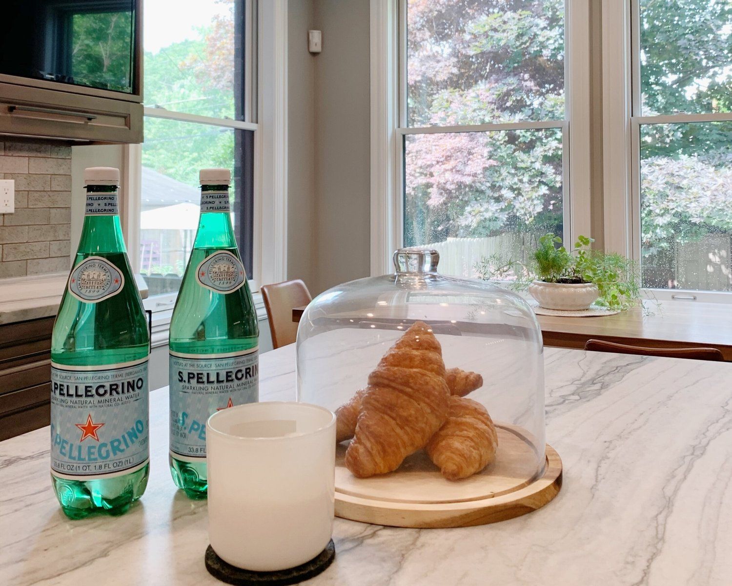 Two bottles of sparkling water, a white candle, and croissants under a glass dome on a marble kitchen counter.