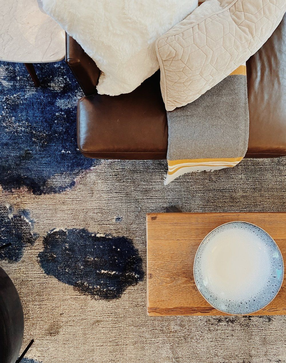 Top-down view of a brown leather sofa with beige pillows and a throw blanket on a patterned blue-and-gray rug with a tray.