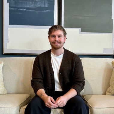 A person with a beard, wearing a white shirt and brown cardigan, sits on a beige sofa in front of abstract wall art.