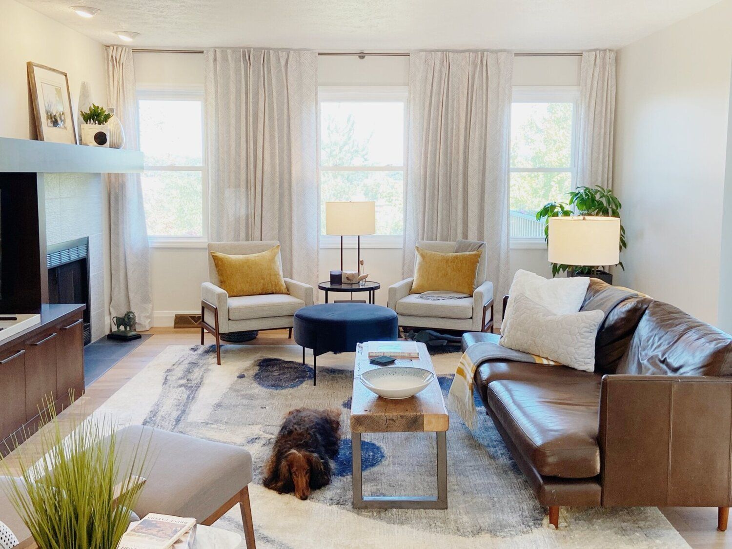 A cozy living room with a brown leather sofa, two light armchairs, a blue ottoman, and a dog resting on a patterned rug.