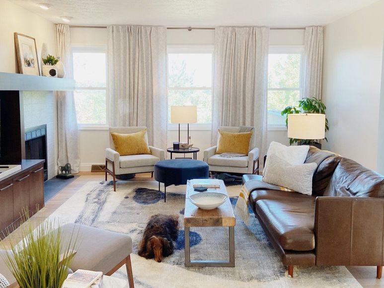A cozy living room with a leather sofa, two light armchairs with yellow pillows, a blue ottoman, and a dog on the rug.