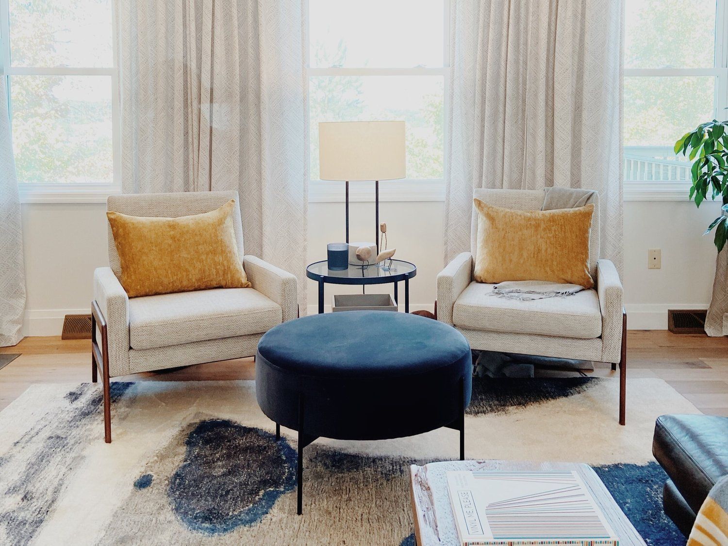 Two light-colored armchairs with yellow throw pillows flank a small table with a lamp, centered before curtains.