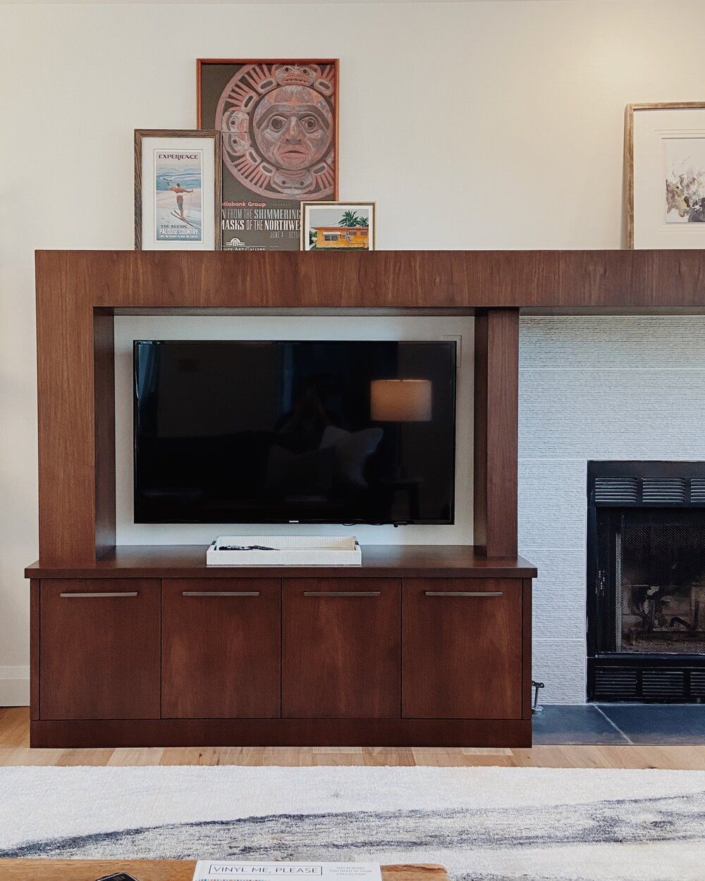 A dark wood cabinet holds a wall-mounted TV beside a brick fireplace, with art displayed on the top shelf.