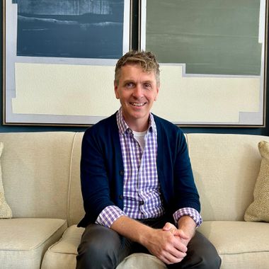 A smiling person in a blue cardigan and plaid shirt sits on a light-colored sofa in front of abstract framed wall art.