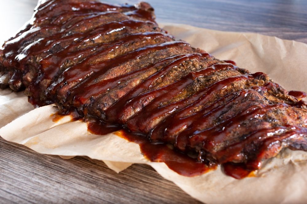 A full rack of smoked barbecue ribs generously glazed with sauce, resting on parchment paper on a wooden surface.