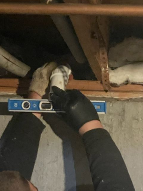 Person in black gloves using a level to align a white plumbing fitting in a basement.
