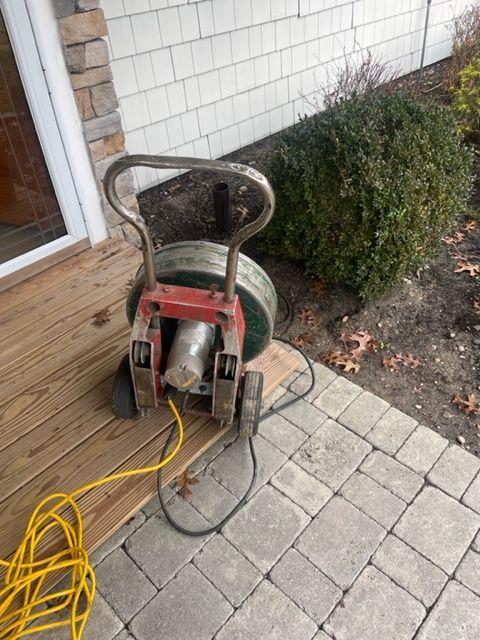 A drain cleaning machine sits on a porch and brick walkway, with a yellow extension cord.