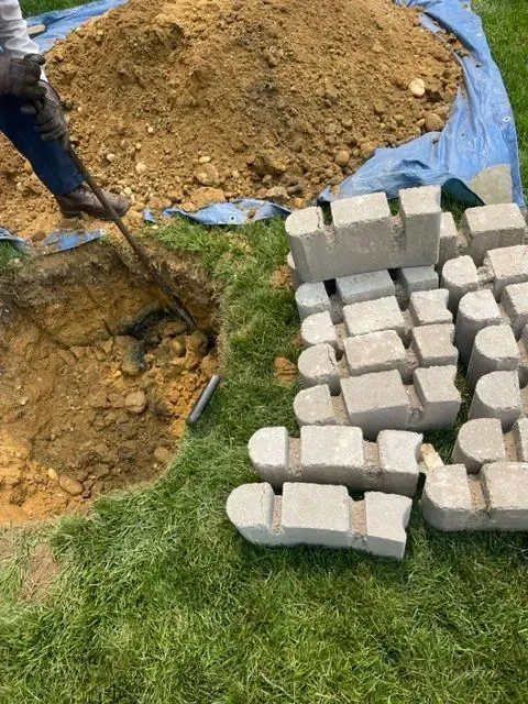 Person digging in a hole near a pile of dirt and a stack of concrete blocks on grass.