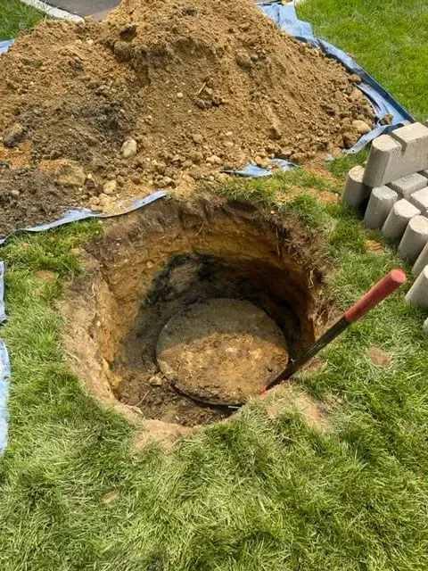 Hole dug in grass, with dirt pile, round concrete lid, and shovel.
