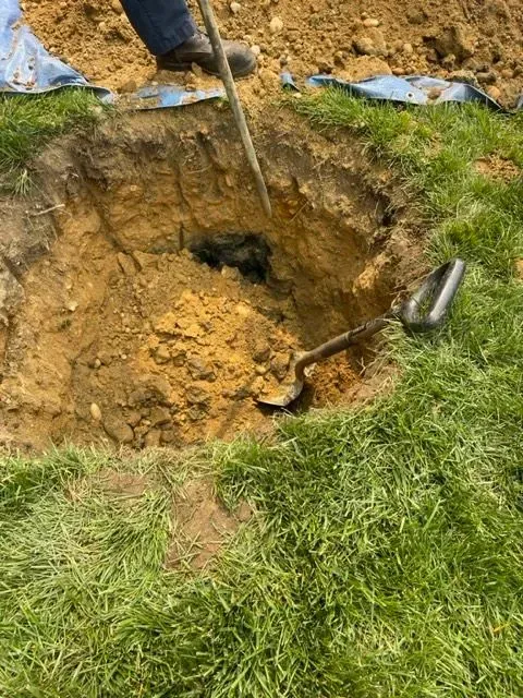 Person digging a deep, dark hole in brown soil, shovel in the hole, with grass in the foreground.