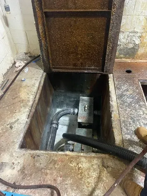 Open metal hatch reveals concrete pit with pipes and equipment; vacuum hose in view.