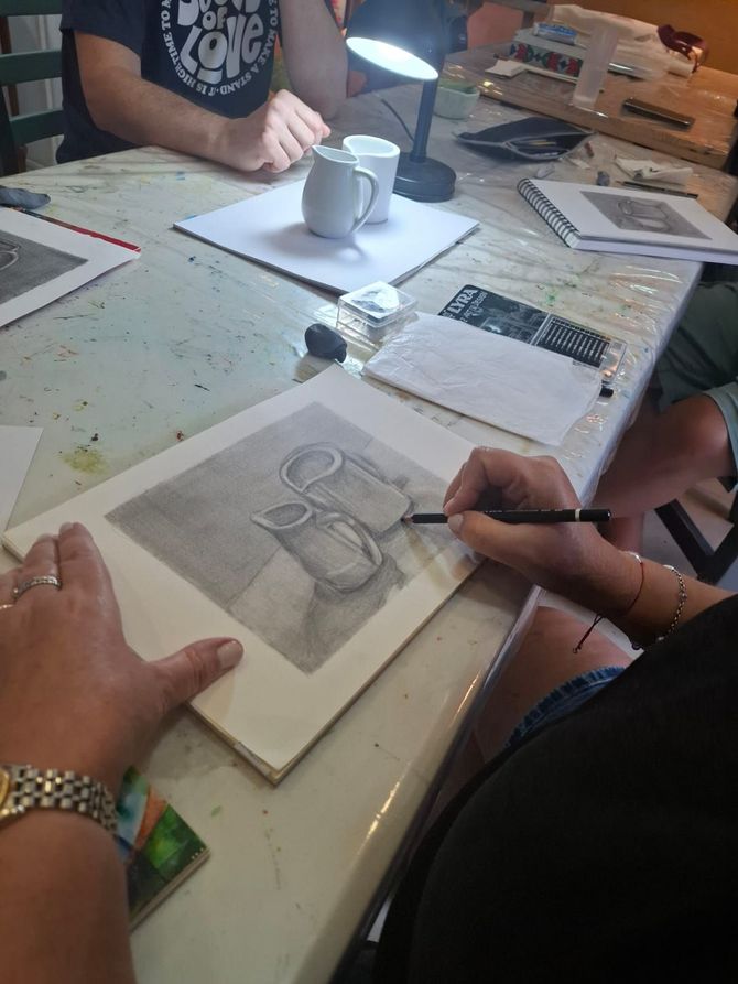 Una persona dibuja una naturaleza muerta de dos jarras blancas en un taller, utilizando un lápiz sobre papel.