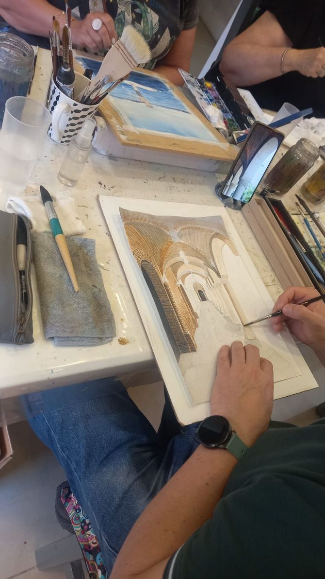Una persona pinta una acuarela detallada de un edificio de piedra sobre papel, sentada a una mesa con otros artistas.