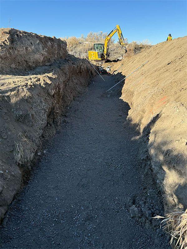A deep trench filled with gravel, yellow excavator in the background, construction site.