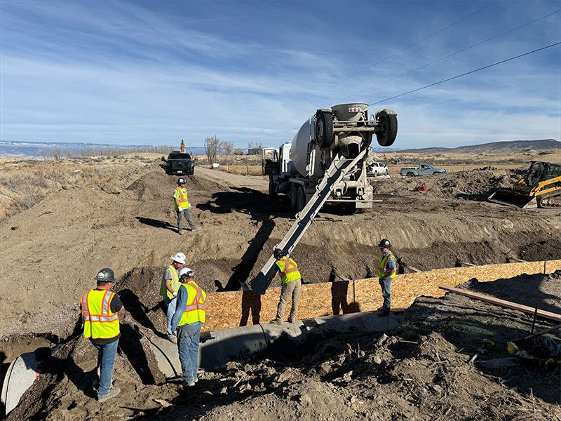 Construction workers pouring concrete from a truck into a trench on a job site under a blue sky.