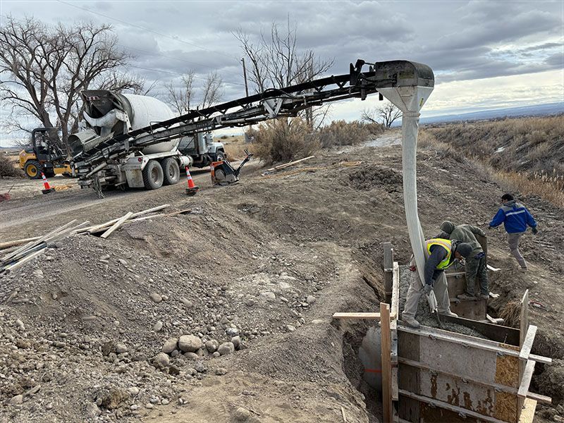 Concrete truck pouring concrete into a construction form on a dirt embankment. Workers are present.