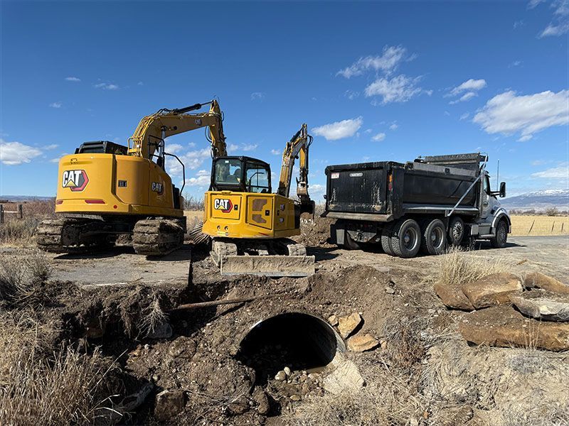 Yellow excavators and dump truck by a road, working on a culvert under blue sky.