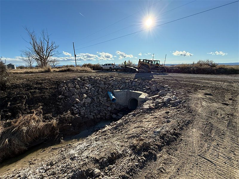 A small creek flows under a road, culvert surrounded by rocks, sunny blue sky with construction equipment.