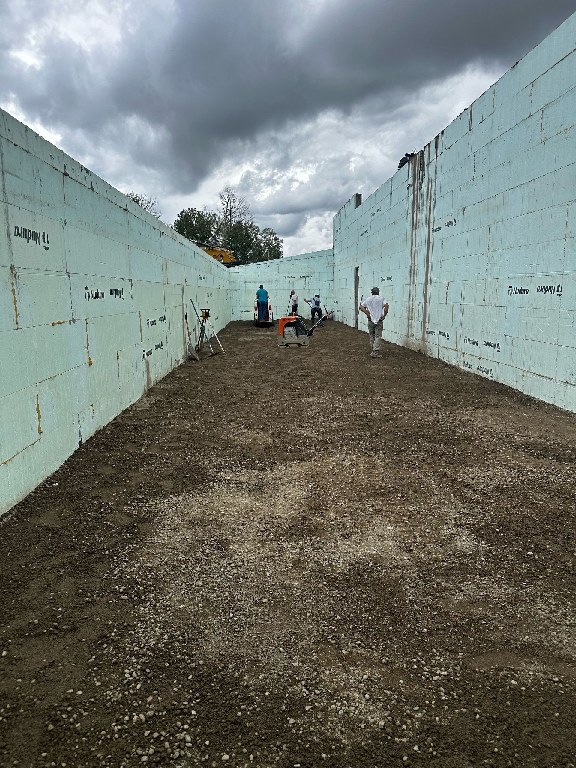 Construction site: Workers inside a walled foundation, spreading gravel on the ground. Cloudy sky.