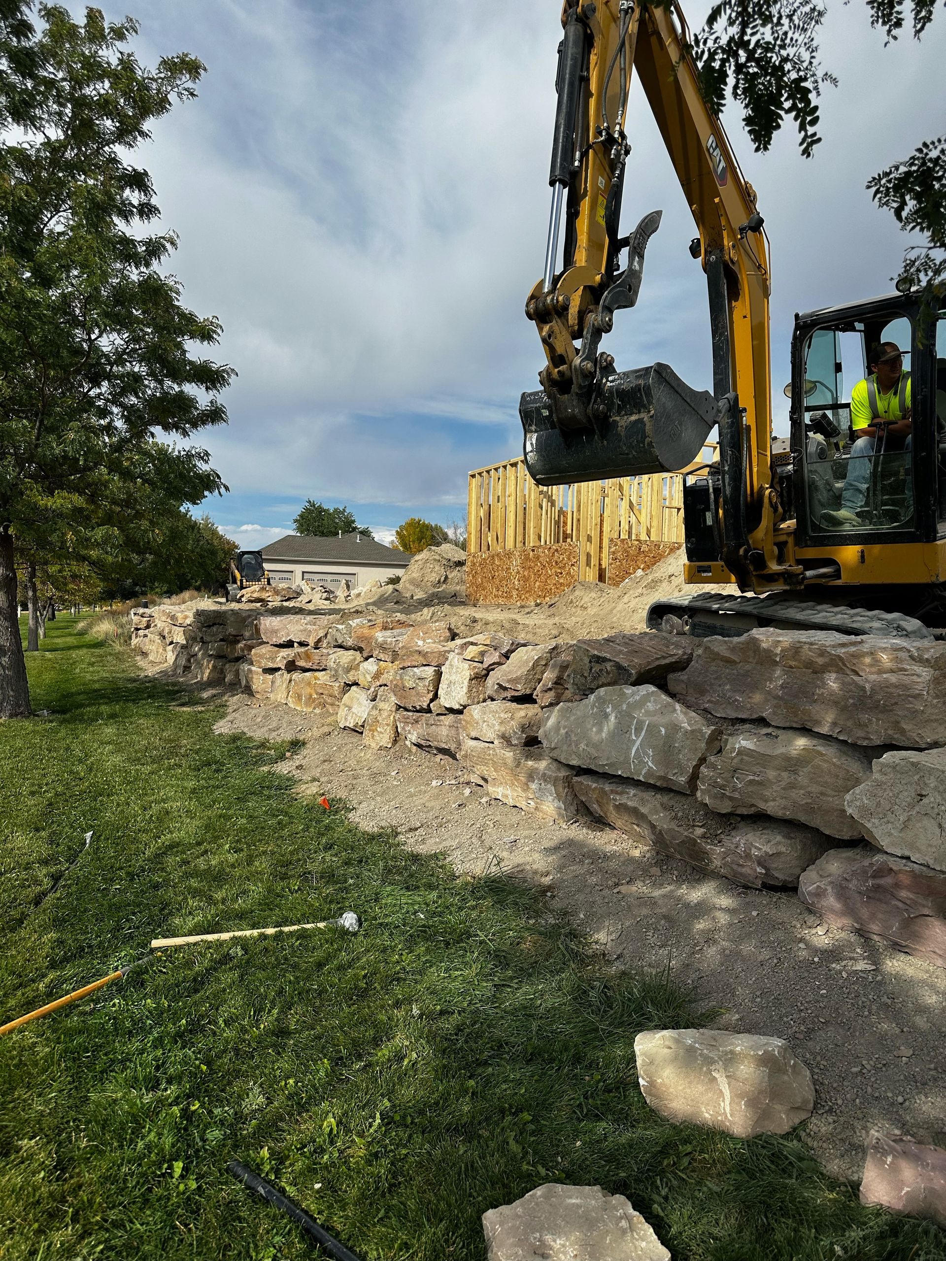 Excavator demolishing a rock wall and wooden structure in a grassy area, under a cloudy sky.