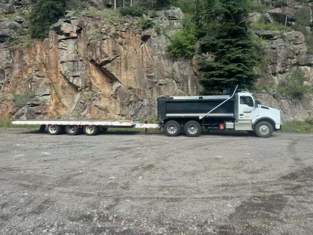 Truck with a flatbed trailer parked on gravel in front of a rocky cliff face.