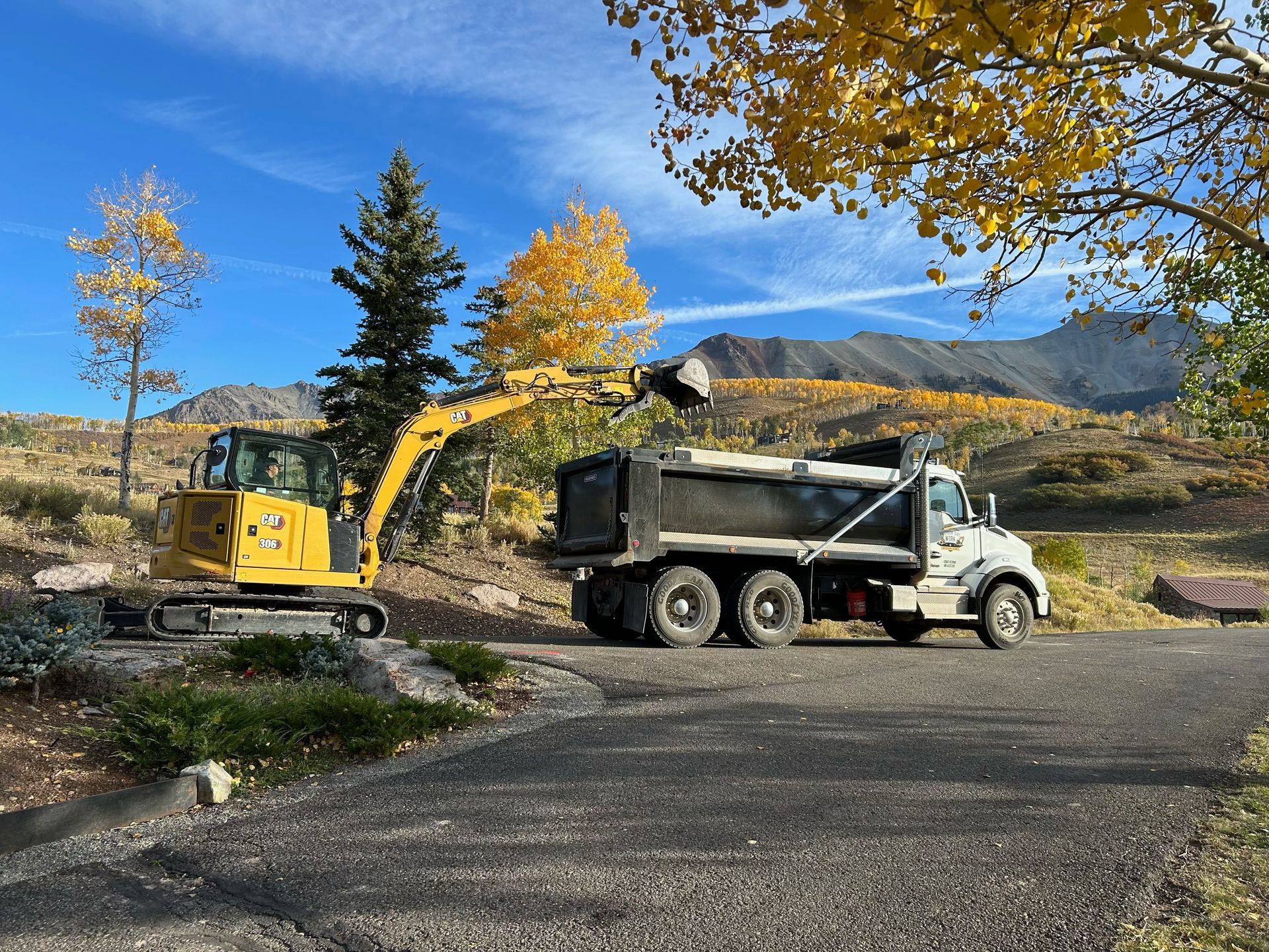 Yellow excavator loading a dump truck on a paved road, with mountains and fall foliage in the background.