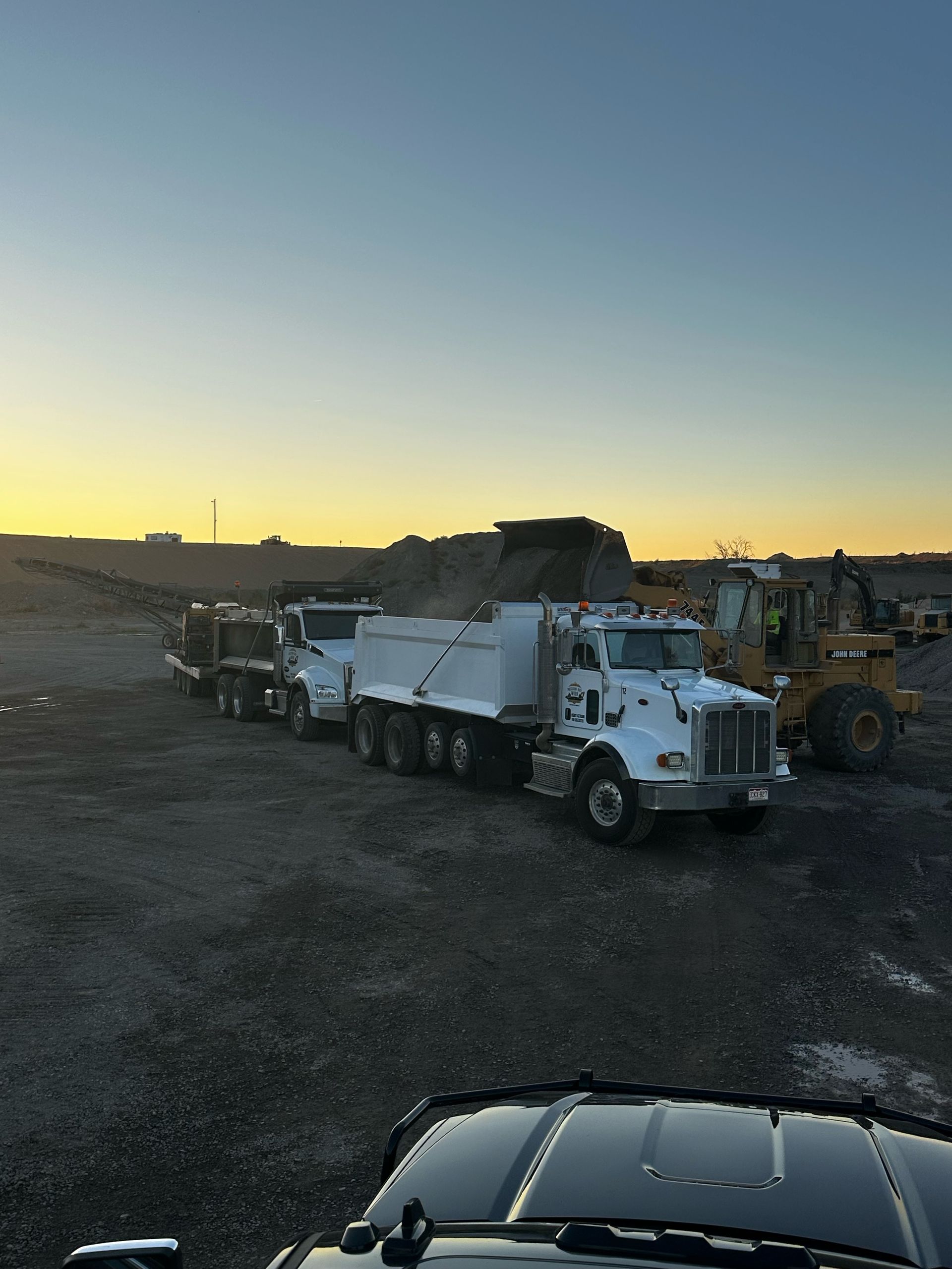 Three white dump trucks lined up, with a loader, at a gravel pit under a dusky sky.