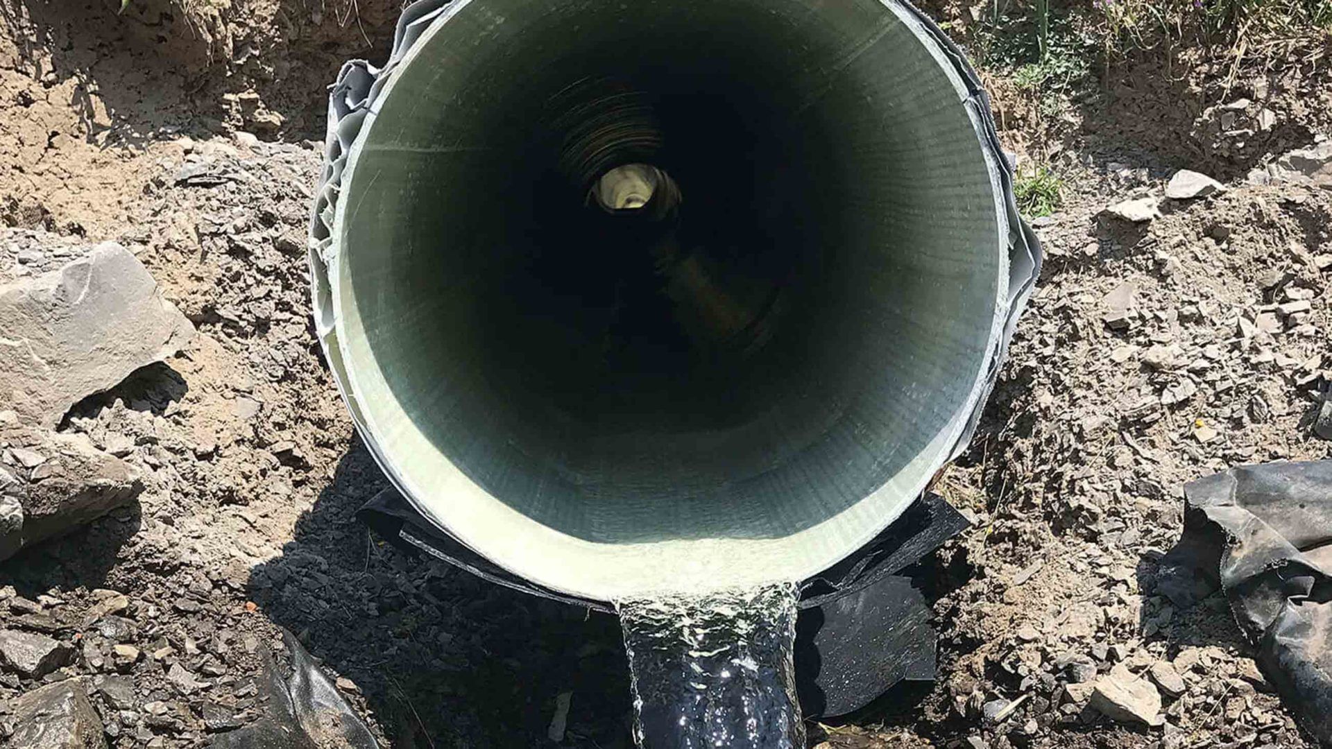 Inside view of a large, black and green pipe; open end facing upward in a dirt trench.