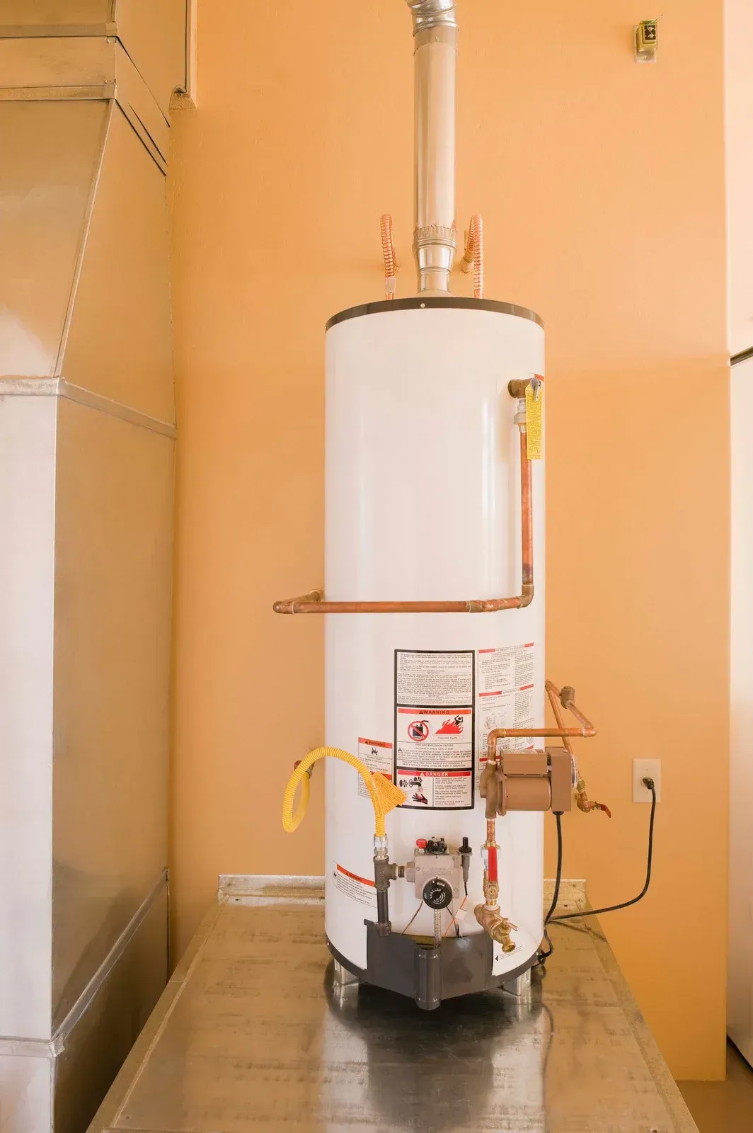 White water heater with copper pipes, vent, and electrical cord on a stainless steel surface against an orange wall.
