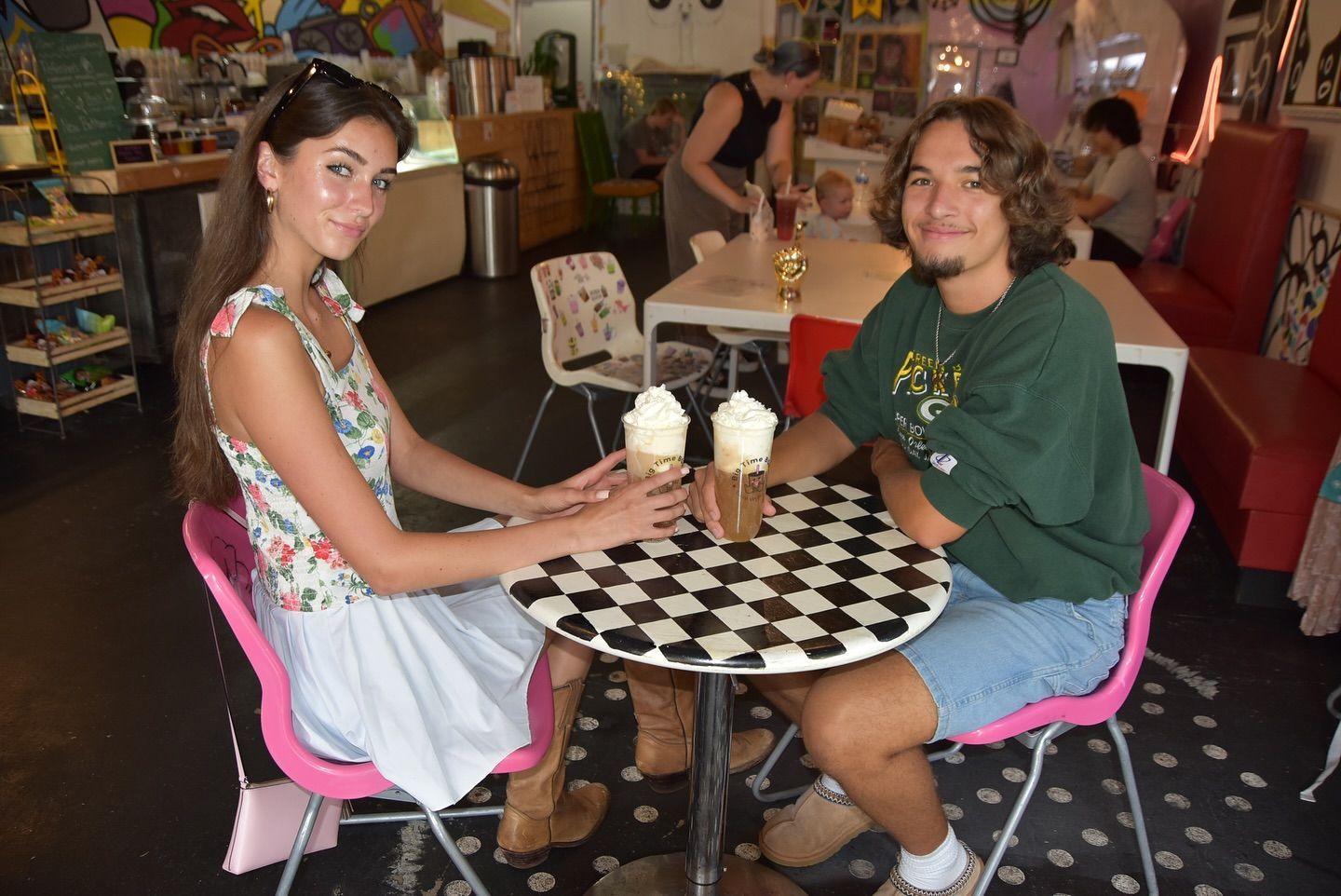 A couple at a checkered table with drinks. Woman in floral top, man in green shirt, pink chairs, cafe setting.