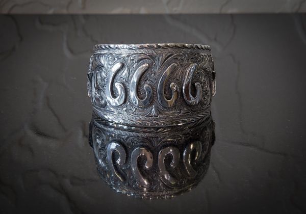 Silver, ornate cuff bracelet on a reflective surface, displaying swirling patterns.