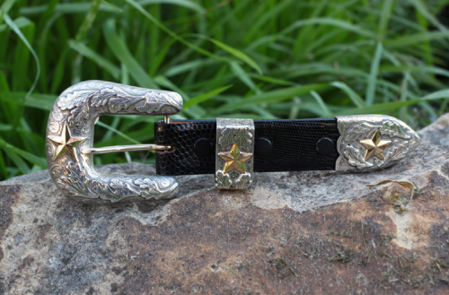 Western-style belt buckle with silver and gold star details, sitting on a rock in front of grass.