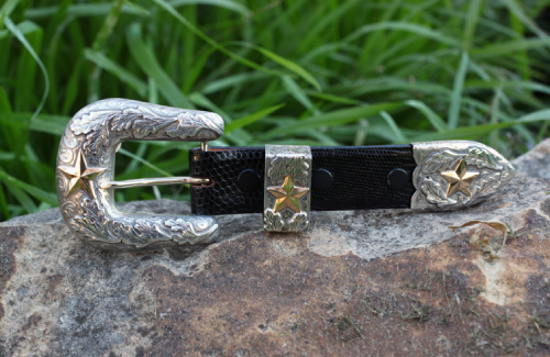 Silver and gold belt buckle with a black leather strap and star accents on a rock.