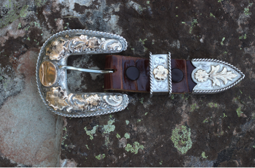 Western style belt with intricate silver and gold buckle set against a textured rock surface.