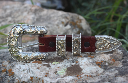 Silver and gold-toned western belt buckle on brown leather strap, set against a rocky background.