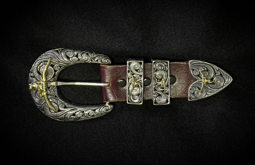 Silver and gold engraved western belt buckle with brown leather strap, on a black background.