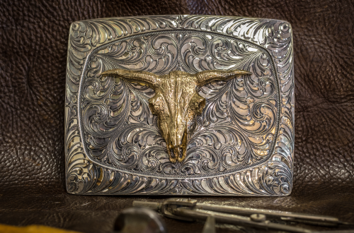 Silver belt buckle with engraved design and gold steer skull in the center.
