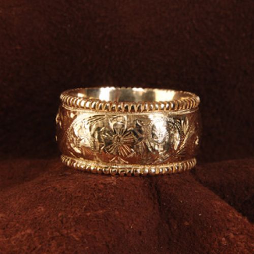 Gold band ring with floral carvings and beaded edges, set on a dark brown surface.