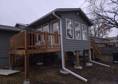 Grey house addition with deck and windows, on concrete supports, with white gutters, outdoors.