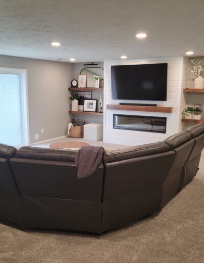 L-shaped dark leather sectional in a finished basement with fireplace, TV, and floating shelves.