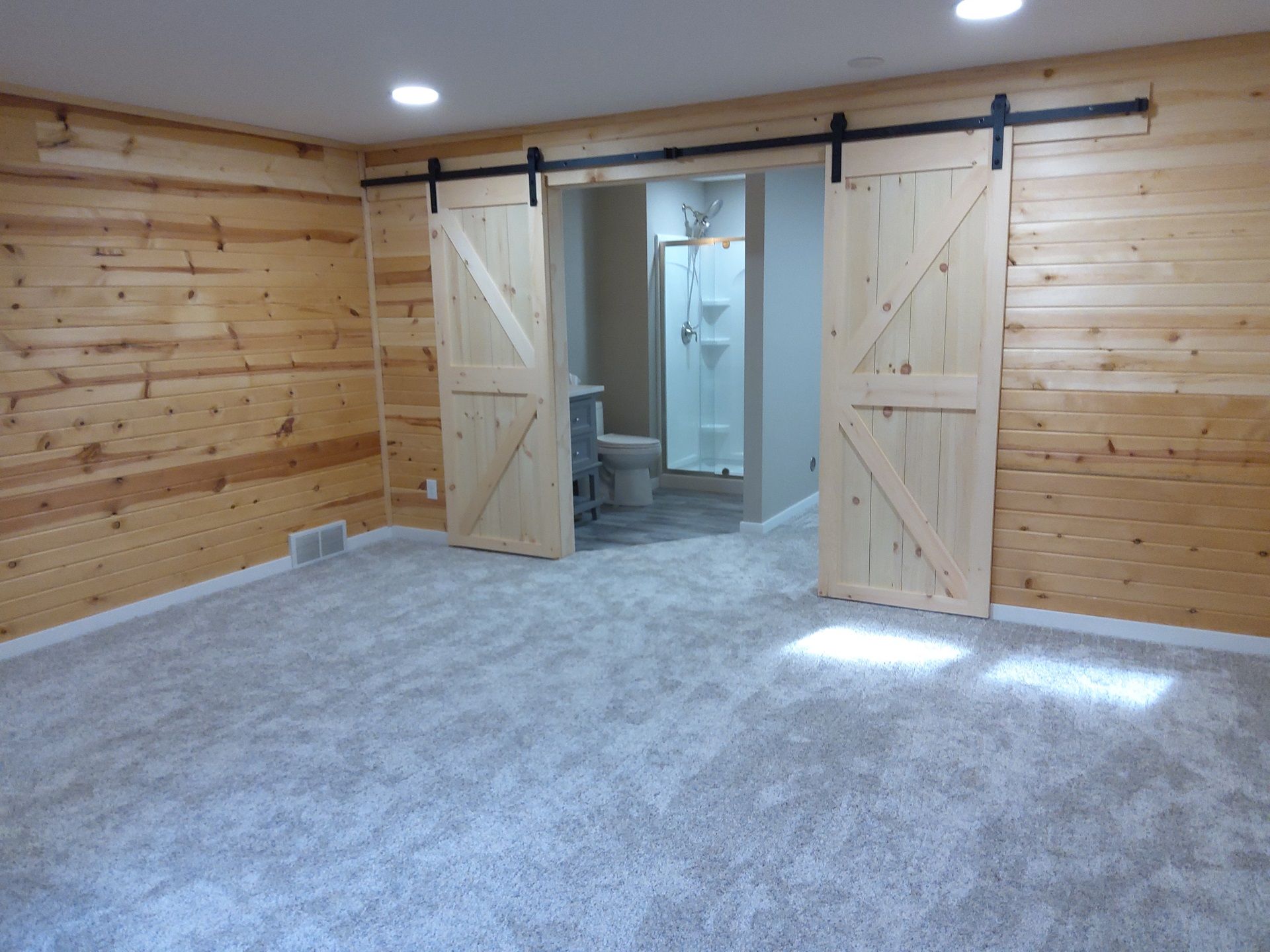 Bedroom with light wood-paneled walls, gray carpet, and two light wood barn doors opening to a bathroom.