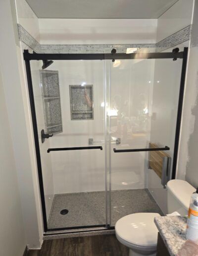 Bathroom with a glass shower enclosure with black framing and sliding doors. The shower has white walls, a gray floor, and a toilet.