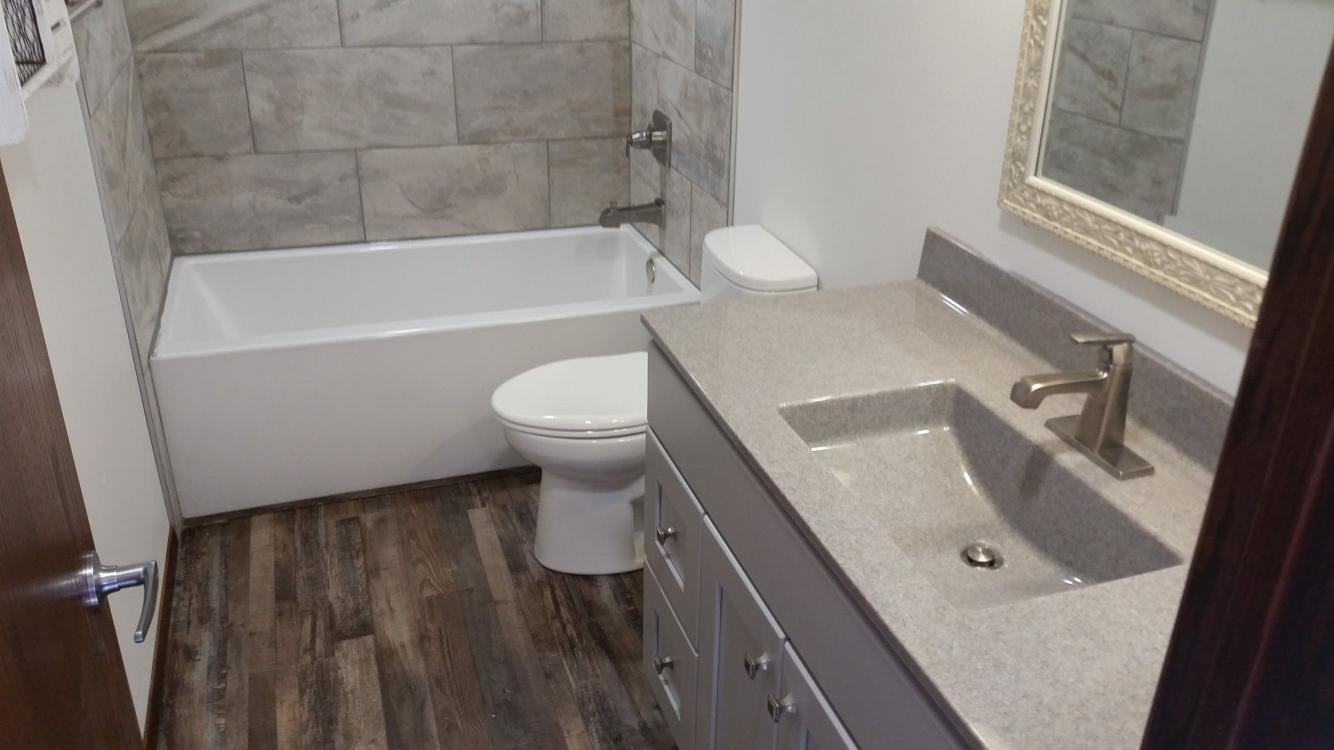 Bathroom with tub, toilet, and vanity with stone-look countertop, dark wood-look flooring, and gray tile shower.
