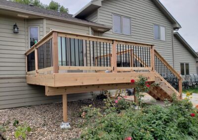 Wooden deck attached to a house with black railing and steps; rose bushes in the foreground.