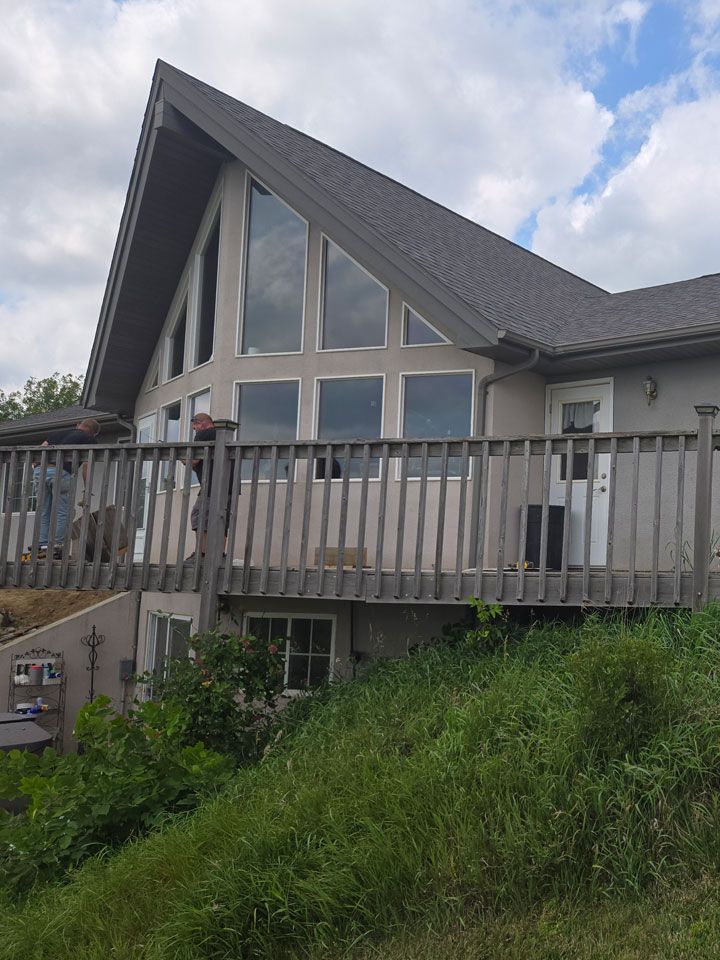 Tan house with a deck and large windows under a gray roof. Green foliage in the foreground.