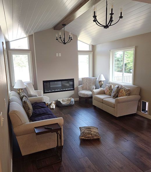Living room with fireplace, sofas, two chandeliers, hardwood floor, and large windows.