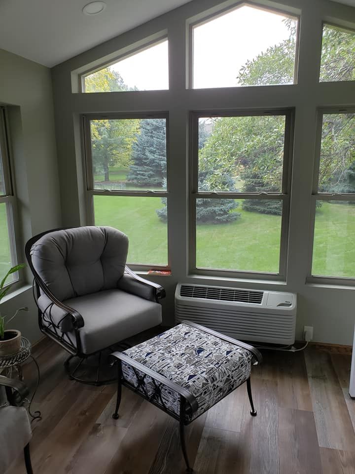 Sunroom with chair and footstool, large windows overlooking a green lawn. Gray furniture, brown floor.