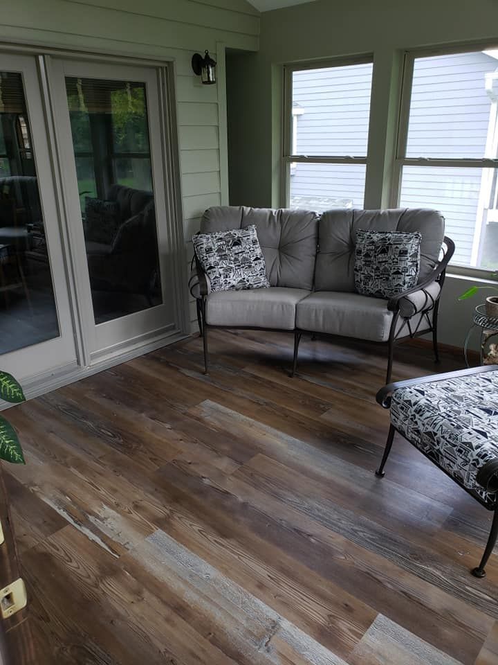 Sunroom with wood-look flooring, gray loveseat with patterned pillows, and a patterned ottoman. Sliding glass doors and windows.