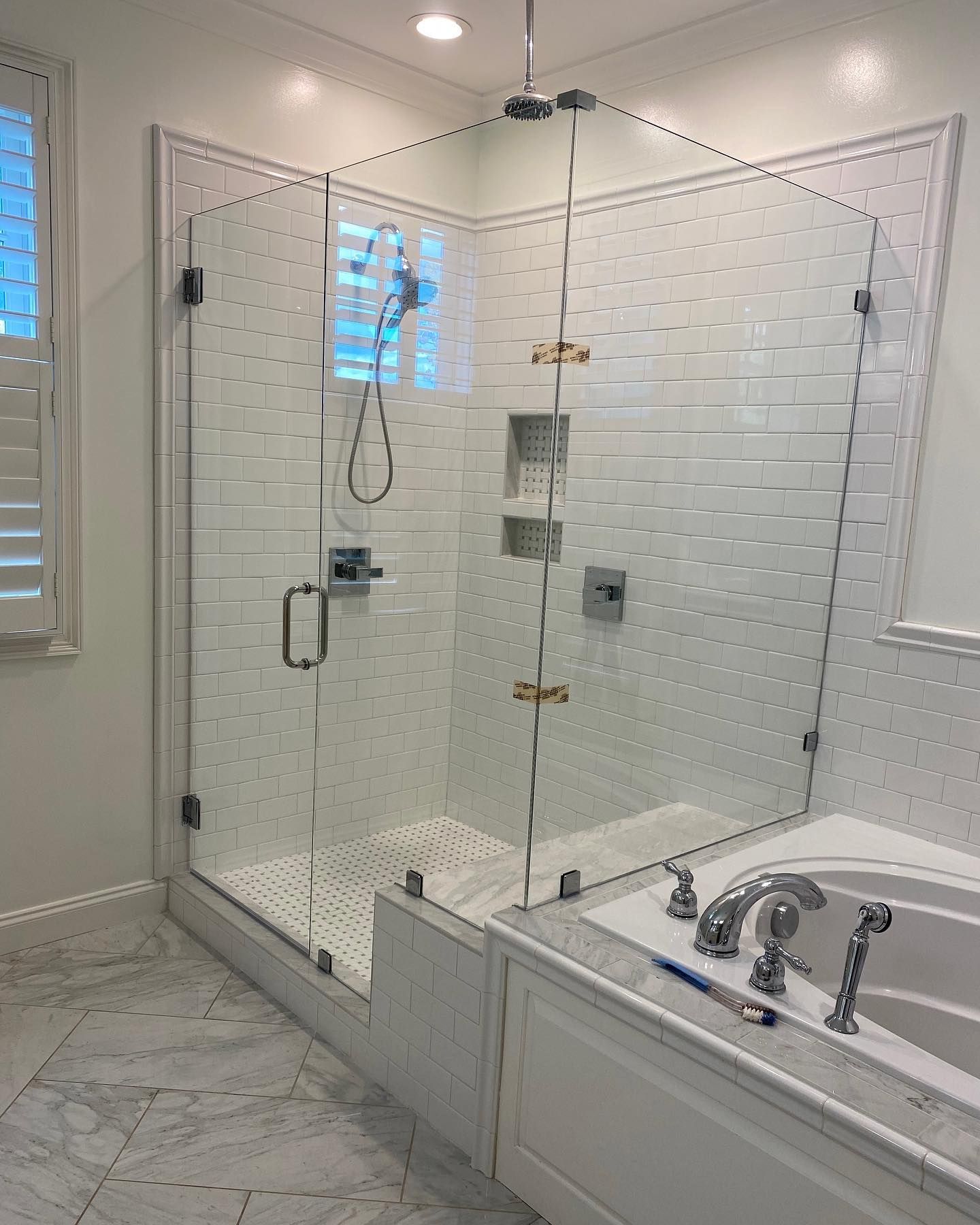 Glass shower enclosure in a white-tiled bathroom with a bathtub. Glass shower enclosure in a white-tiled bathroom with a bathtub.