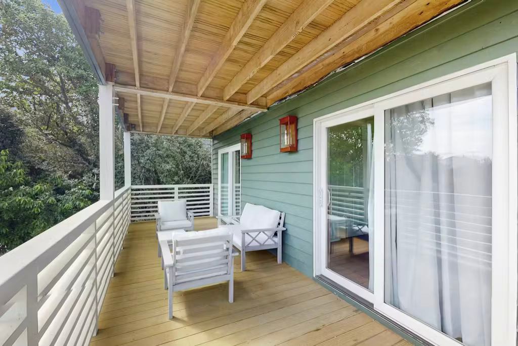 Balcony with white furniture, wood flooring, and green siding. Trees in the background, sliding glass door. Balcony with white furniture, wood flooring, and green siding. Trees in the background, sliding glass door.